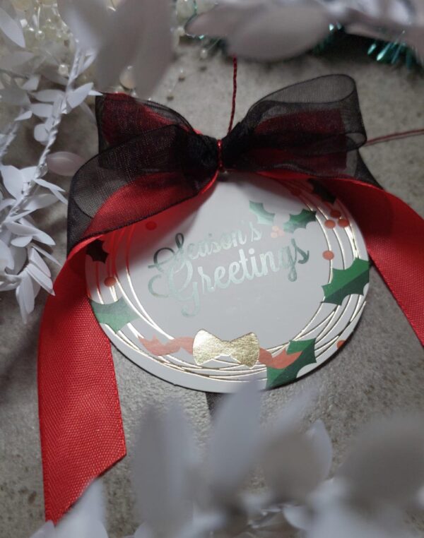 Circular Christmas tag with Seasons Greeting foiled in gold and a red and black ribbon tied to it.